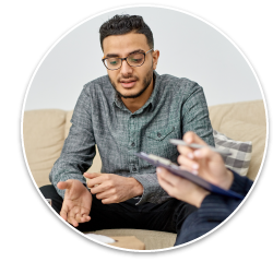 A South-Asian man in his late 20's/early 30's sitting on a couch, having a discussion with a counselor holding a clipboard.