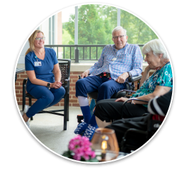 A white woman caregiver, wearing a blue top and an employee ID badge, sits on a porch talking to an elderly white man and an elderly white woman in a wheelchair.