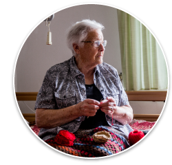 An elderly white woman sits on her bed white she knits a colorful blanket.