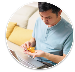 An Asian man in his 30's with a laptop resting on his lap, taking one white pill out of an orange prescription pill bottle.